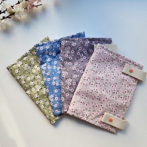 May include: Four fabric pouches in shades of green, blue, gray, and pink, each adorned with a delicate floral pattern. The pink pouch features two beige straps with orange button closures. The pouches are arranged in a fan shape, showcasing their colorful designs.