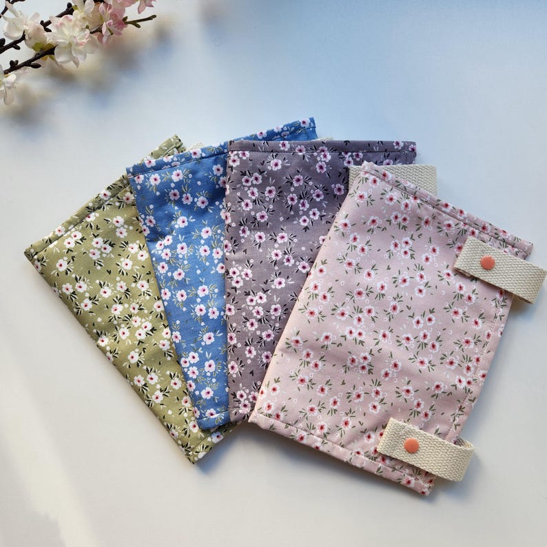 May include: Four fabric pouches in shades of green, blue, gray, and pink, each adorned with a delicate floral pattern. The pink pouch features two beige straps with orange button closures. The pouches are arranged in a fan shape, showcasing their colorful designs.