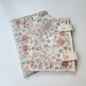 May include: Two floral fabric items with a cream background, featuring pink, white, and blue flowers and green leaves. Each item has a cream-colored border and cream straps with pink buttons. The items are likely fabric organizers or covers.