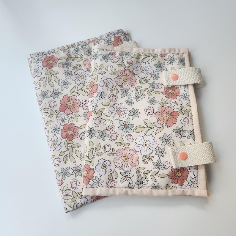 May include: Two floral fabric items with a cream background, featuring pink, white, and blue flowers and green leaves. Each item has a cream-colored border and cream straps with pink buttons. The items are likely fabric organizers or covers.