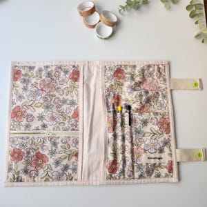 May include: A floral fabric journal cover with multiple pockets and a zipper closure. The cover features a cream background with pink, red, and green flowers. The text "AtelierHippie" is visible on a small tag. Several rolls of decorative tape are in the background.