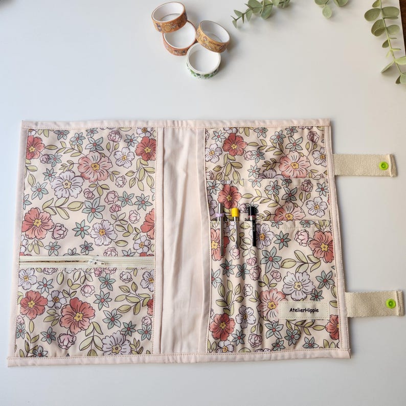 May include: A floral fabric journal cover with multiple pockets and a zipper closure. The cover features a cream background with pink, red, and green flowers. The text "AtelierHippie" is visible on a small tag. Several rolls of decorative tape are in the background.