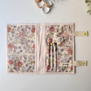 May include: Floral fabric pen case with a zipper pocket and multiple pen slots. The case features a cream background with pink, red, and green floral patterns. The case has a beige strap with a green button closure. The text "AtelierHippie" is visible.