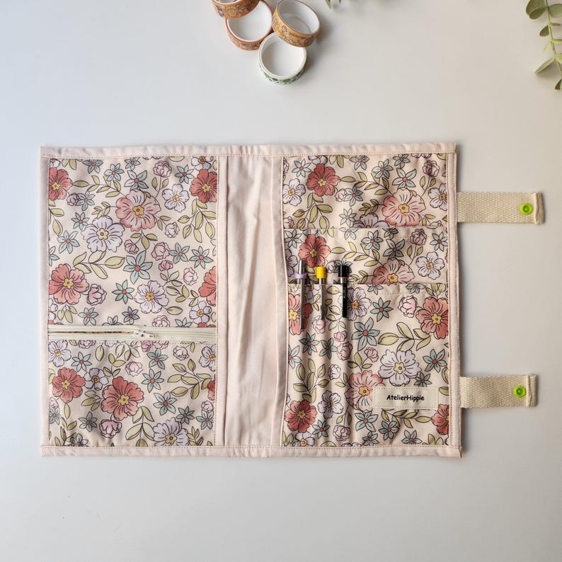 May include: Floral fabric pen case with a zipper pocket and multiple pen slots. The case features a cream background with pink, red, and green floral patterns. The case has a beige strap with a green button closure. The text "AtelierHippie" is visible.