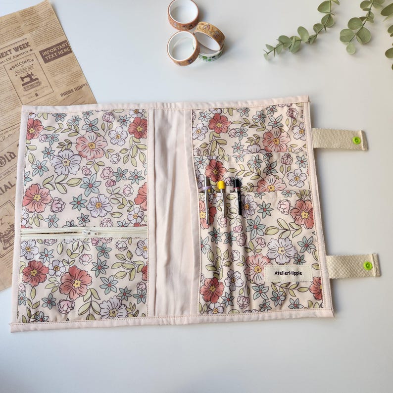May include: A floral print pen case with a light pink background. The case features a zipper pocket, pen holders, and a snap closure. The floral pattern includes pink, white, and blue flowers with green leaves. The text "Atelier Hippie" is visible.