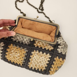 May include: A handmade crochet purse in a granny square pattern. The bag features a dark gray, beige, and yellow color scheme. It has a metal clasp closure and a chain strap. The interior is lined with a tan fabric.