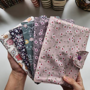 May include: A collection of fabric-covered journals and notebooks with floral patterns. The covers feature various colors, including pink, purple, and blue, with a button closure on the front. The notebooks are held in a person's hands.