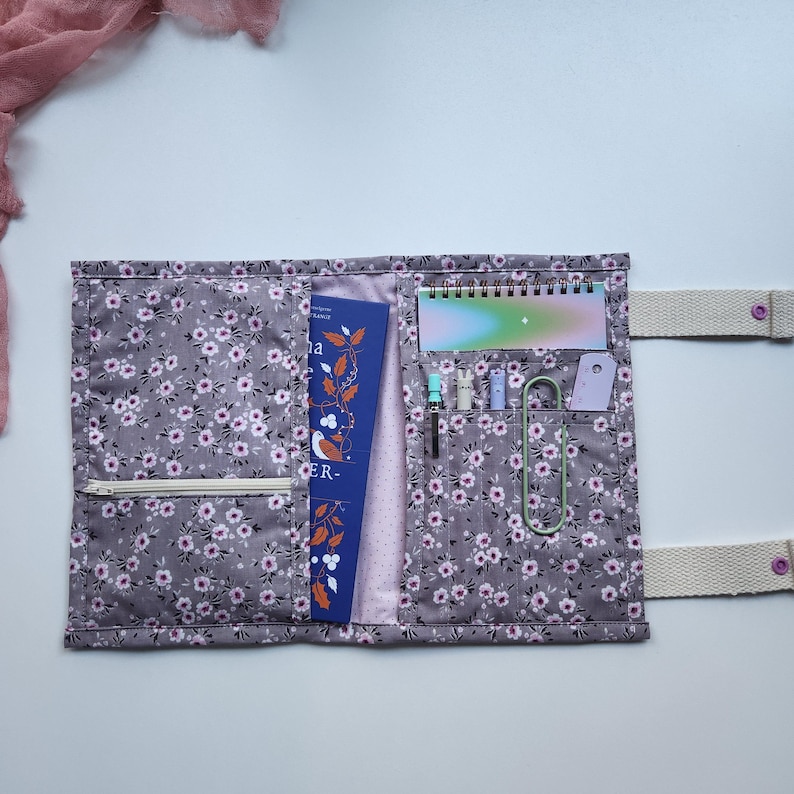 May include: A gray floral fabric book cover with a zipper pocket, holding a book, notepad, pens, and paper clips. The cover has a light pink floral pattern and a cream-colored strap with a pink button closure.