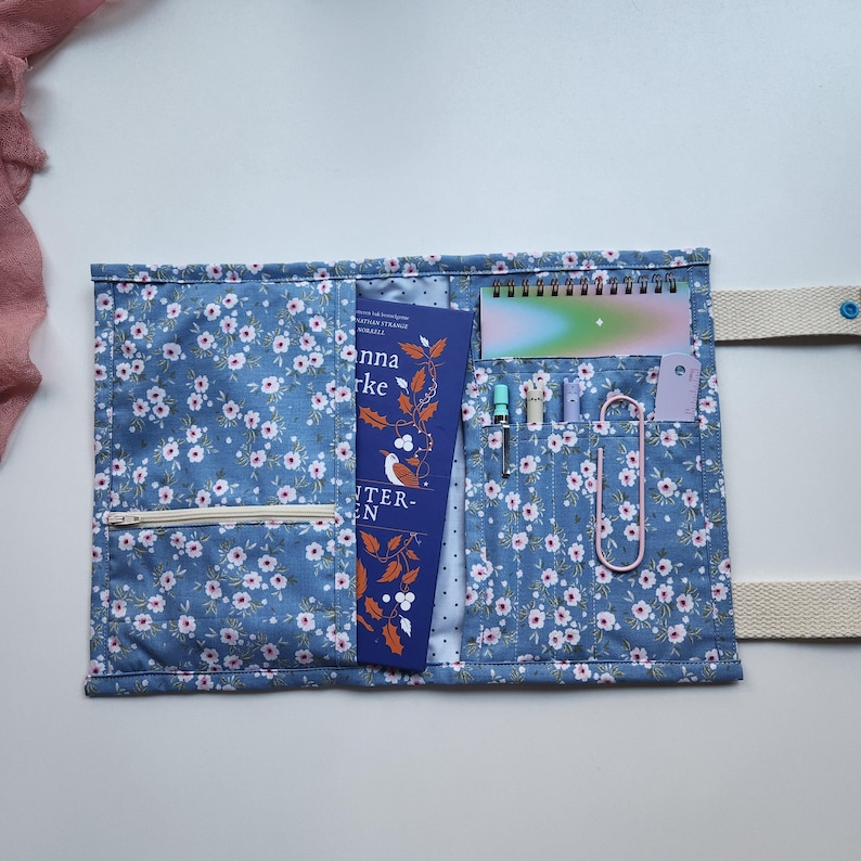 May include: Blue floral fabric book sleeve with multiple pockets. The sleeve holds a book, a small notebook, pens, and a paperclip. A zipper pocket is visible on the front. The book has the title "Anna Burke" on the cover.