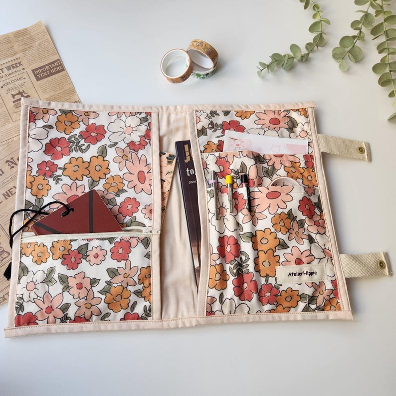 May include: Open floral print organizer with multiple pockets. The fabric features a pattern of orange, peach, and red flowers on a cream background. The organizer has a zippered pocket, pen holders, and a strap closure. The brand name "Atelier Hippie" is visible.