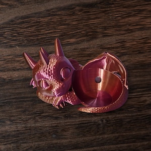 May include: A metallic purple dragon-shaped container with a lid. The dragon has a detailed, textured surface and large, round eyes. The container is open, revealing a circular interior. The object is set on a dark wood surface.
