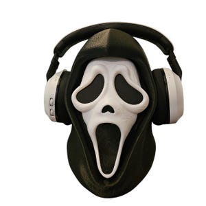 May include: A white Scream mask with black eye sockets and an open mouth, wearing white and black headphones. The mask is set against a black hood, creating a spooky and modern design.