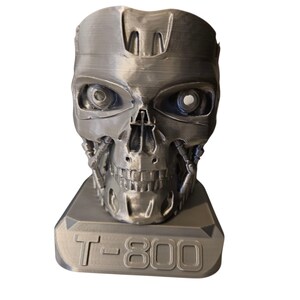 May include: A silver-coloured sculpture of a Terminator T-800 skull. The sculpture has detailed mechanical features, including visible eye sockets and a jaw. The base of the sculpture is rectangular and features the text "T-800".