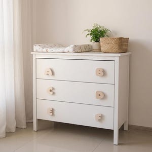 Puede incluir: Una cómoda blanca de tres cajones con pomos de madera clara en forma de letras A, B, C, D, E y F. Sobre ella hay un cambiador con estampado, una pequeña planta verde y una cesta de mimbre.