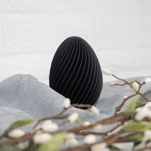Peut inclure: Un objet décoratif noir en forme d'œuf avec un motif en spirale nervuré. L'objet repose sur un tissu gris, avec une branche de fleurs blanches au premier plan. L'arrière-plan est un mur de briques blanches.