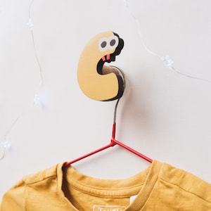 May include: A whimsical, yellow and black cartoon character-shaped wall hook with a red metal hanger. The hook is mounted on a white wall, with a yellow t-shirt hanging from the hanger.