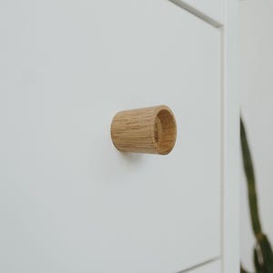 Puede incluir: Primer plano de un tirador de armario cilíndrico de madera clara, montado en una puerta de armario blanca. El tirador tiene un centro hueco y presenta vetas de madera natural.