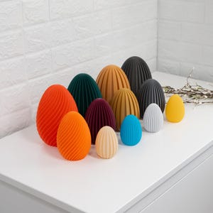 Op de afbeelding: Een verzameling decoratieve, eivormige objecten in verschillende kleuren, waaronder oranje, groen, geel en blauw. De eieren hebben een gestructureerd, geribbeld ontwerp en zijn op een wit oppervlak geplaatst. Thema: Pasen of lente decoratie.