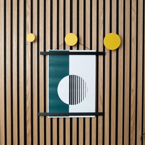 Puede incluir: Un póster enmarcado en negro con un diseño geométrico verde y blanco cuelga de una pared de madera con tres ganchos de pared circulares amarillos.
