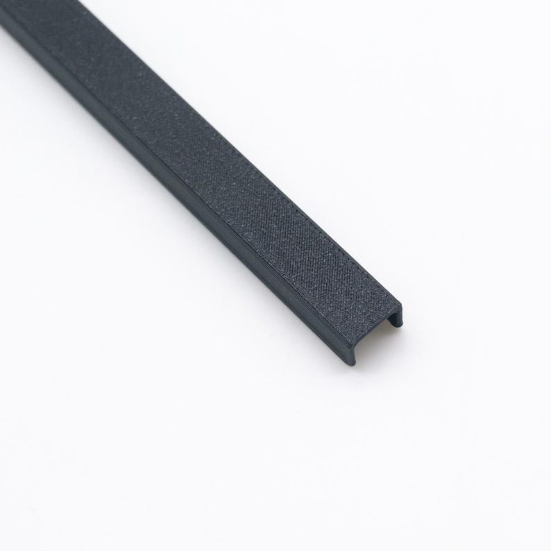 Peut inclure: Une longue bande de canal en forme de U, &eacute;troite, avec une finition mate textur&eacute;e gris fonc&eacute;. L'objet pr&eacute;sente un profil lin&eacute;aire coh&eacute;rent avec des extr&eacute;mit&eacute;s ouvertes, montrant une structure rigide et une texture uniforme sur toute sa longueur.