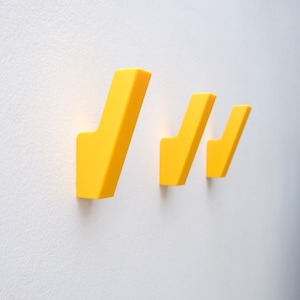 May include: Three bright yellow wall hooks mounted on a white textured wall. The hooks are in a modern, minimalist style, with a simple, angular design. The hooks are of varying sizes.