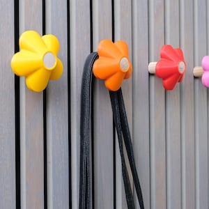 May include: A row of flower-shaped wall hooks in yellow, orange, red, and pink. The hooks are mounted on a gray wooden slatted wall. A black strap hangs from the orange hook. The hooks have a wooden center.