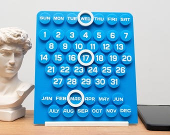 May include: A blue perpetual desk calendar with white numbers and text. The days of the week and months are printed at the top and bottom. The current date is highlighted with a white ring. A bust of a statue is in the background.
