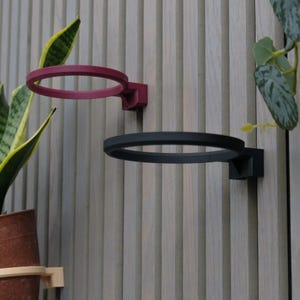 May include: Two wall-mounted plant holders. One is a deep red, the other is black. Each has a circular ring and a square mounting bracket. The holders are mounted on a light wood-paneled wall. Green plants are visible in the background.