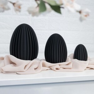 May include: Three black decorative egg sculptures of varying sizes, each with vertical ridged detailing. They rest on a light peach-colored fabric against a white background. The largest egg is approximately 8 inches tall.