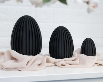May include: Three matte black egg-shaped sculptures of varying sizes, each featuring a vertical ribbed texture. The objects are arranged in a row and rest upon a draped, light beige fabric that gathers around their bases.