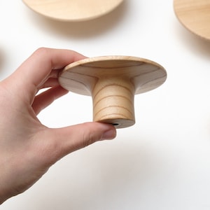 May include: A wooden knob with a wide, flat top and a cylindrical base. The knob is made of light-coloured wood with a grain pattern.