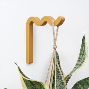 Puede incluir: Un gancho de pared color amarillo mostaza con un borde superior ondulado y serpenteante. El gancho está montado en una superficie plana y sostiene una planta colgante mediante un cordel de yute fino de color natural con hojas verdes.