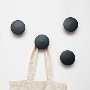 May include: Four black wooden wall hooks with a conical design are mounted on a white wall. A beige tote bag hangs from one of the hooks. The hooks have a matte finish and a minimalist aesthetic.