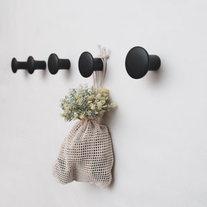 May include: Five circular black wooden wall hooks mounted in a horizontal row. The hooks feature a smooth, rounded front face and a cylindrical base. A beige mesh drawstring bag containing small green and yellow artificial flowers hangs from one hook.