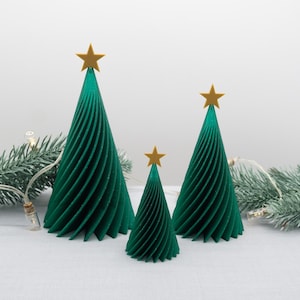 May include: Three green spiral Christmas trees of different sizes, each with a gold star on top. The trees are set on a white surface, with fairy lights and evergreen branches in the background. A festive holiday decoration.