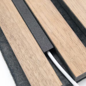 Könnte beinhalten: Nahaufnahme einer hölzernen Ladestation mit einem dunkelgrauen Metallakzent und einem weißen Kabel. Die Holzlatten sind hellbraun und auf einer dunkelgrauen Filzbasis angeordnet. Das Kabel kommt aus der Ladestation.