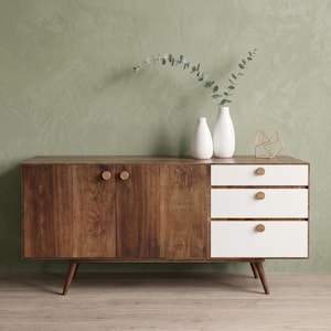Puede incluir: Un aparador de madera con dos puertas de armario con pomos redondos de madera y tres cajones blancos con pomos redondos a juego. El mueble se apoya sobre cuatro patas cónicas de madera y está decorado con dos jarrones blancos y una estructura de alambre.