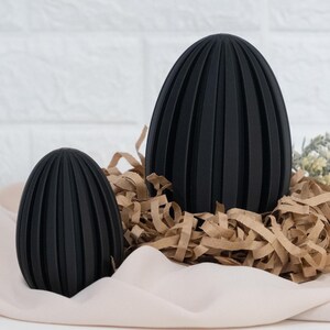 Op de afbeelding: Twee zwarte decoratieve eieren met verticale geribbelde details. De eieren liggen in bruine papiersnippers op een lichtroze stof. De achtergrond toont een witte bakstenen muur en een paar groene takjes.