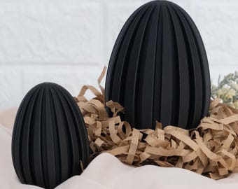 May include: Two matte black egg-shaped objects with vertical ribbed textures. The objects are positioned upright, with one larger than the other, featuring a consistent dark finish and a series of parallel ridges running from the top to the base.