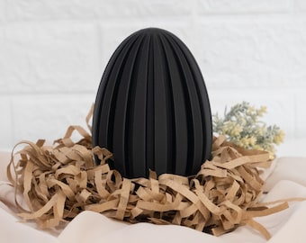 May include: A black, egg-shaped decorative object with vertical grooves, resting in a bed of brown shredded paper. The egg is the focal point, set against a white brick wall and a cream-colored fabric.