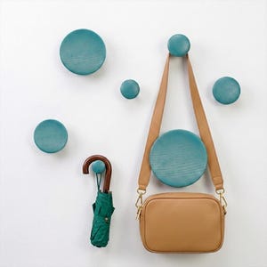 May include: A set of six circular teal wooden wall hooks of varying sizes mounted on a wall. A tan leather crossbody bag hangs from one of the larger hooks, and a folded green umbrella with a curved wooden handle hangs from a smaller hook.