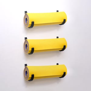 Op de afbeelding: Drie opgerolde gele yogamatten met paarse randen zijn aan een witte muur bevestigd. Elke mat wordt op zijn plaats gehouden door twee zwarte metalen beugels. De matten zijn verticaal gelijkmatig verdeeld, wat een strakke en georganiseerde uitstraling creëert.