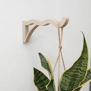 May include: A beige wall-mounted plant bracket with a wavy, curved top edge and a triangular support brace. A natural jute twine hanger is looped over the end of the bracket, supporting a green and yellow variegated snake plant leaf structure.