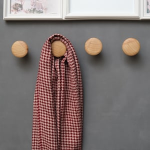Op de afbeelding: Drie ronde houten wandhaken zijn bevestigd aan een grijze muur. Een bordeauxrode en crèmekleurige sjaal met pied-de-poule patroon hangt aan een van de haken. De haken zijn gelijkmatig verdeeld en hebben een natuurlijke houtnerf. Boven de haken bevindt zich een wit omkaderde foto.