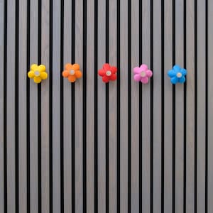 May include: Five colorful flower-shaped wall hooks in yellow, orange, red, pink, and blue are mounted on a vertical striped wooden panel. Each hook has a white center, adding contrast to the vibrant petals.