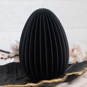 May include: A black, egg-shaped decorative object with vertical ribbed detailing. The object sits on a black leaf-shaped tray with gold trim, against a white background with floral accents. The design is modern and minimalist.