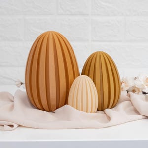 May include: Three decorative Easter eggs in a variety of sizes. The largest egg is a deep brown colour with vertical ridges. The middle egg is a lighter brown colour with vertical ridges. The smallest egg is a light cream colour with vertical ridges. The eggs are sitting on a light brown fabric.