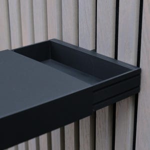 Puede incluir: Estante rectangular negro montado en la pared con una bandeja empotrada. El estante está montado en una pared de madera clara con listones verticales. El estante tiene un diseño minimalista.