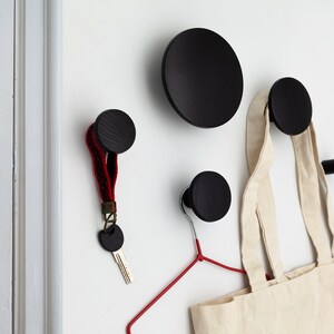 Op de afbeelding: Vier zwarte houten wandhaken, twee ronde en twee paddenstoelvormige, zijn gemonteerd op een witte muur. Een rode hanger is bevestigd aan een van de ronde haken. Een witte draagtas hangt aan een van de paddenstoelvormige haken. Een sleutel met een rood-zwarte gevlochten band is bevestigd aan de andere paddenstoelvormige haak.
