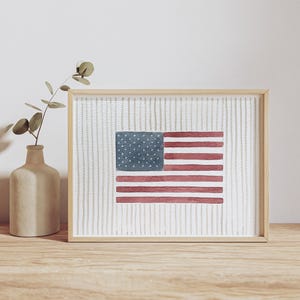 May include: A framed watercolor print featuring the United States flag centered on a white background with vertical grey dashed lines. The flag displays a blue canton with white dots and horizontal red stripes. A light wood frame surrounds the artwork.